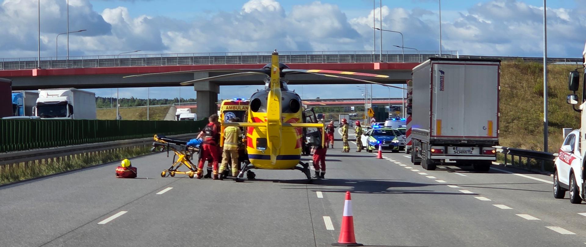 Widok na autostradę z zamkniętym ruchem. Na jezdni stoi żółty śmigłowiec ratunkowy z napisem "AMBULANS", wokół którego pracuje kilku ratowników medycznych i strażaków, ustawiając nosze transportowe. W tle wiadukt, samochody ciężarowe na poboczu, radiowóz policyjny z włączonymi światłami oraz pomarańczowy pachołek drogowy na pierwszym planie. Niebo błękitne z dużymi białymi chmurami.