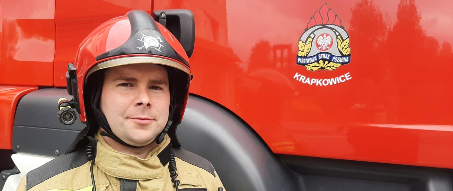 Na tle samochodu pożarniczego stoi strażak w ubraniu specjalnym i czerwonym hełmie. Strażak ukazany jako zdjęcie portretowe - od klatki piersiowej w górę. Na drzwiach pojazdu pożarniczego widoczne logo PSP, a pod nim napis KRAPKOWICE. 