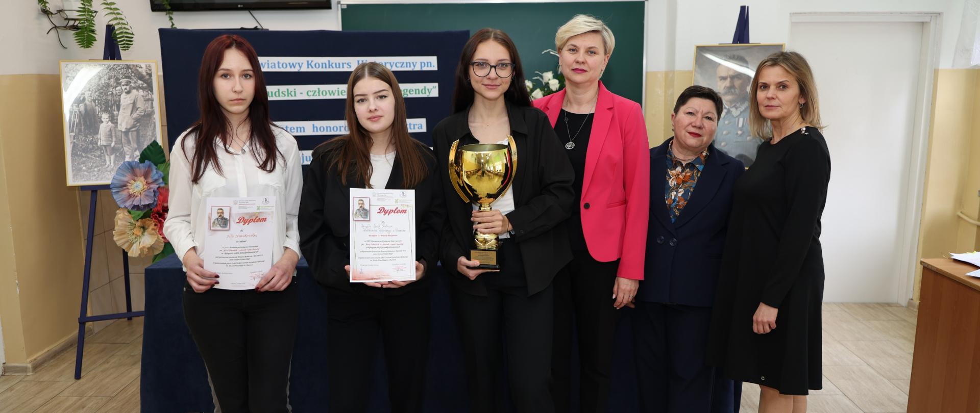Grupa osób pozuje w sali szkolnej na tle tablicy konkursowej; jedna osoba trzyma puchar, a dwie trzymają dyplomy