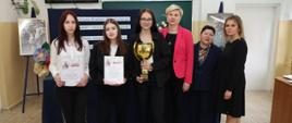 Grupa osób pozuje w sali szkolnej na tle tablicy konkursowej; jedna osoba trzyma puchar, a dwie trzymają dyplomy