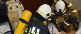 Ćwiczenia w Pakosławiu. Dwóch strażaków udziela pomocy osobie poszkodowanej, która leży na podłodze w jednym z pomieszczeń. Strażacy mają białe hełmy oraz aparaty oddechowe.