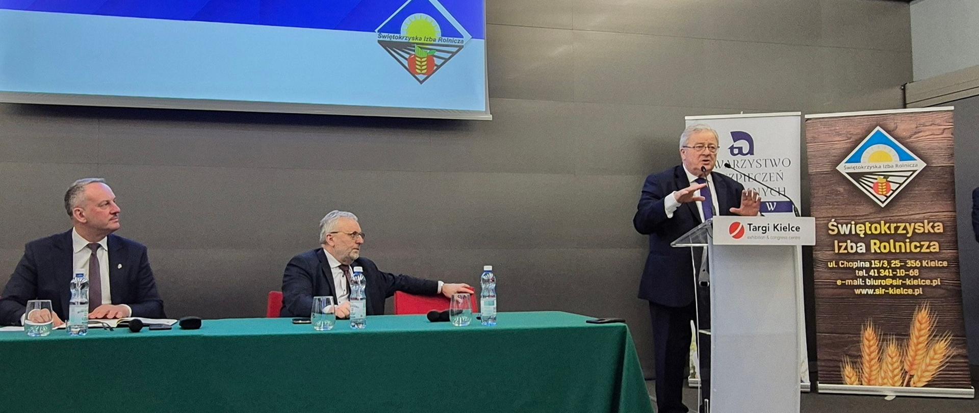 Zdjęcie przedstawia trzech mężczyzn w garniturach. Dwóch spośród nich siedzi przy zielonym stole, jeden zaś przemawia przy mównicy. Za plecami panów jest ekran z napisem Wspólne Posiedzenie Rad Powiatowych Świętokrzyskiej Izby Rolniczej