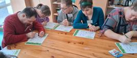 Grupa osób siedzących przy dużym, drewnianym stole w sali warsztatowej. Uczestnicy pracują nad kartami edukacyjnymi ułożonymi przed nimi. Karty zawierają zadania rysunkowe, schematy oraz elementy do kolorowania związane z tematyką zdrowotną, m.in. obrazki przedstawiające rośliny, przedmioty i proste scenki. Przy stole siedzi kilka osób, każda z nich intensywnie pochylona nad swoją kartą pracy, trzymając w dłoniach zielone ołówki. 