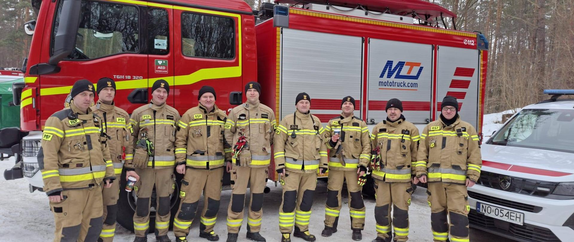 Grupa strażaków w jasnych mundurach na tle samochodu strażackiego. Zima, biało od śniegu.