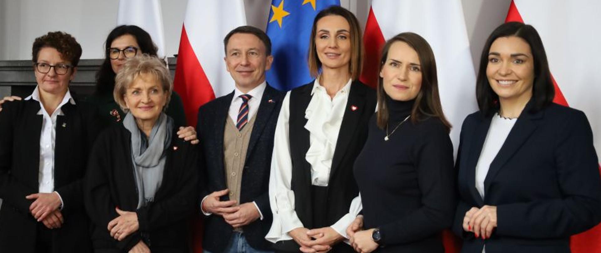 Sala konferencyjna, siedem osób stojących na tle flag polskich i unijnych.