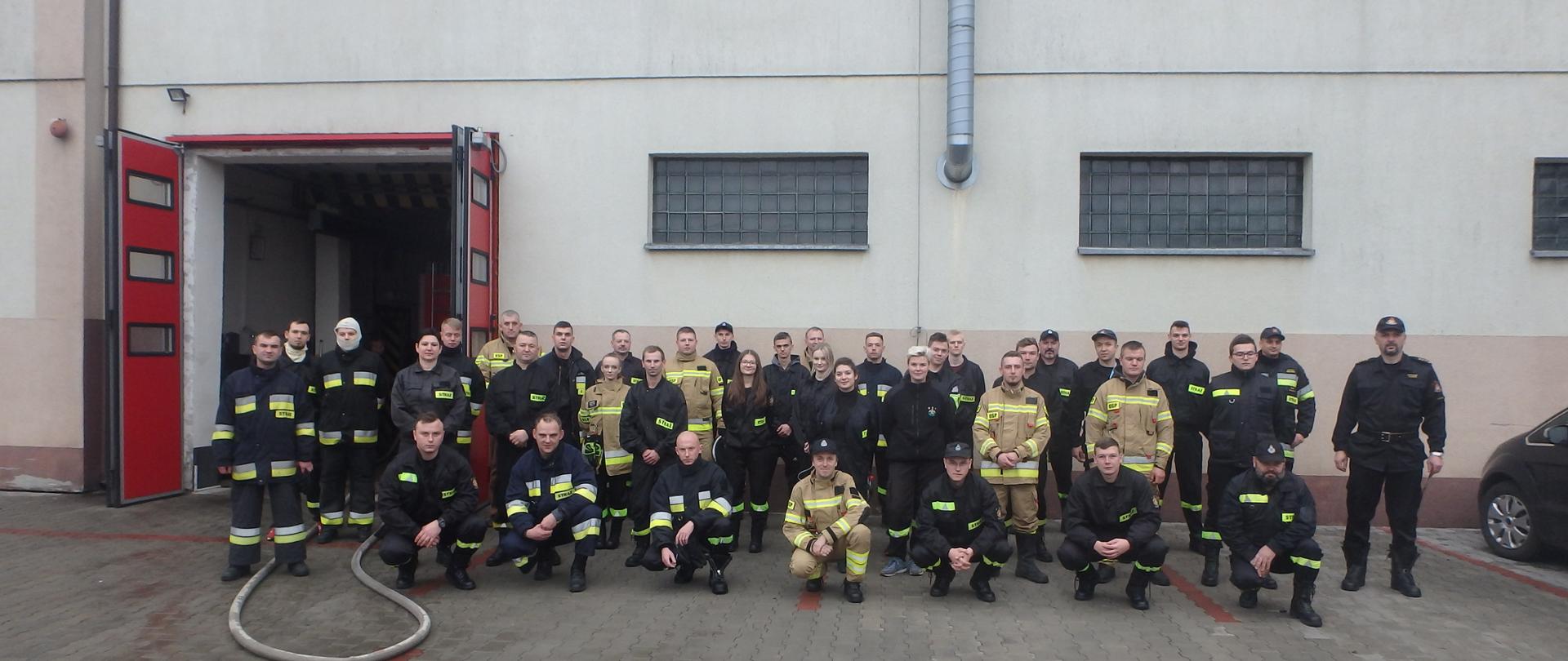 Na placu wewnętrznym komendy grupa strażaków ochotników wraz z komisją egzaminującą robią sobie zdjęcie grupowe.