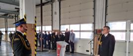 Strażak w mundurze ze sznurem stoi przed pomorskim komendantem wojewódzkim Państwowej Straży Pożarnej. Niedaleko stoją zaproszeni goście.