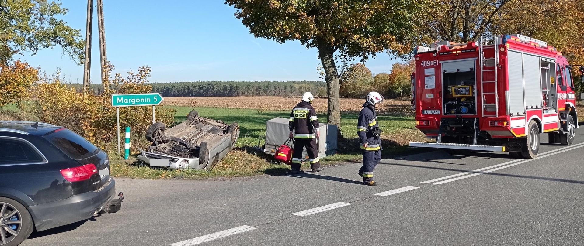 Zdjęcie przedstawia strażaków, którzy usuwają skutki wypadku drogowego.
W tle droga, samochody osobowe uszkodzone, samochód strażacki i pole.
