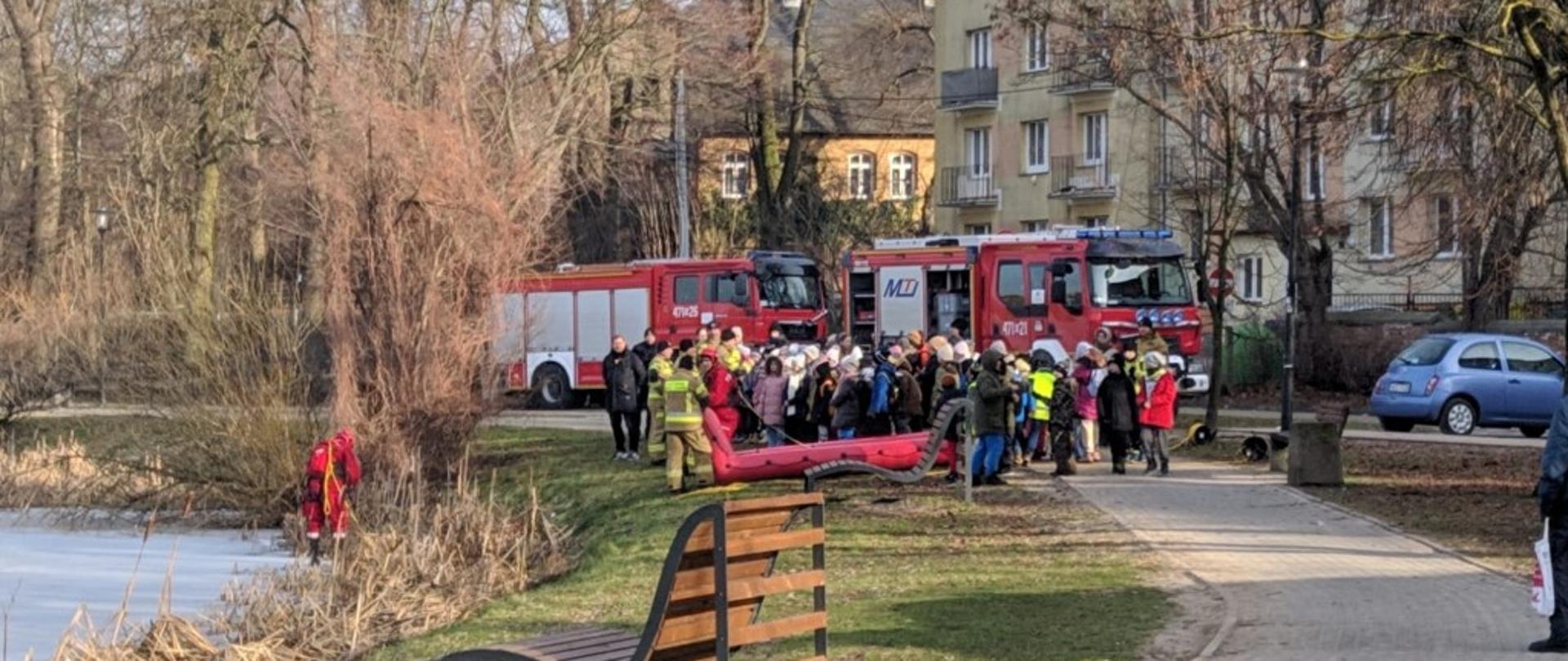 Na zdęciu widoczna grupa strażaków oraz dzieci, sprzęt do ratownictwa lodowego, w tle dwa samochody pożarnicze.