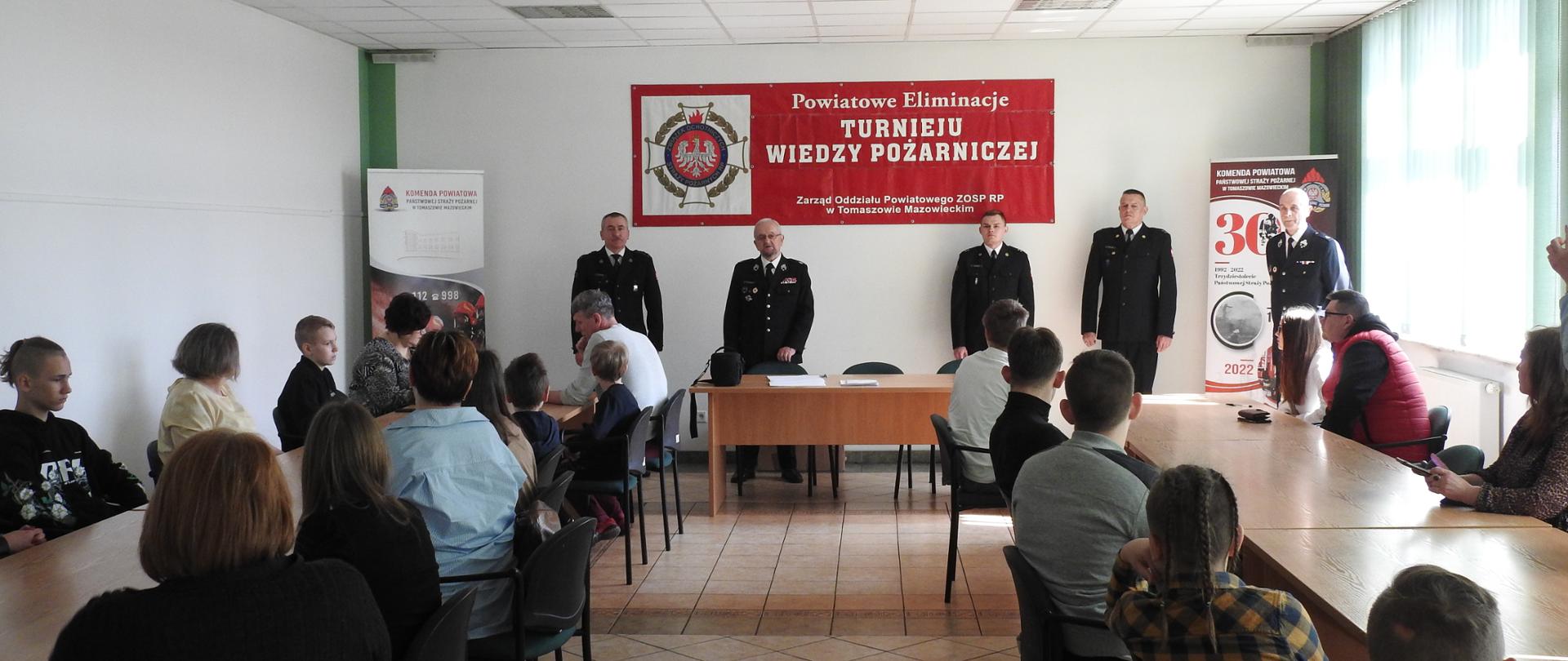 Pięciu strażaków w mundurach wyjściowych stoi na tle jasnej ściany. Nad ich głowami wisi baner z logo Ochotniczych Straży Pożarnych oraz napisem Powiatowe eliminacje Turnieju Wiedzy Pożarniczej. Przed nimi po obu stronach za stołami siedzą dzieci i opiekunowie.