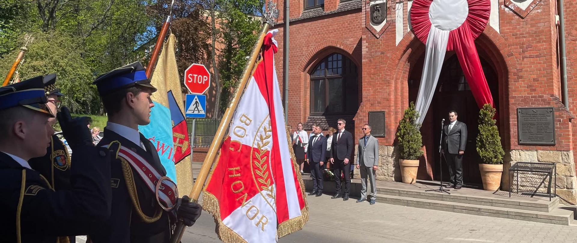 Zdjęcie przedstawia oficjalne obchody rocznicy uchwalenia Konstytucji 3 Maja, odbywające się przed gmachem z czerwonej cegły. Na pierwszym planie widoczny jest poczet sztandarowy złożony z funkcjonariuszy w galowych mundurach, którzy trzymają flagi, w tym biało-czerwoną z napisem „BÓG HONOR OJCZYZNA”. Jeden z mundurowych oddaje honor, salutując w stronę uroczystości. Nad wejściem do budynku umieszczono wielką, biało-czerwoną rozetę z długimi wstęgami, która stanowi centralny element dekoracyjny podkreślający patriotyczny charakter wydarzenia.
W tle, przy wejściu do budynku, stoi grupa mężczyzn w garniturach, z których jeden przemawia do mikrofonu ustawionego na podwyższeniu. Pozostali uczestnicy stoją w postawie zasadniczej, zachowując powagę należną uroczystości. Po bokach wejścia znajdują się krzewy w ozdobnych donicach oraz tablice pamiątkowe wmurowane w elewację. Całość sceny dopełnia słoneczna pogoda, bezchmurne niebo oraz zieleń drzew widoczna za znakiem drogowym, co nadaje obchodom podniosłą i jasną oprawę.