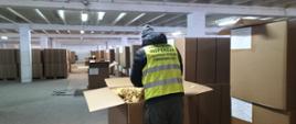 Mężczyzna w żółtej kamizelce z logo PIORiN na plecach na tle dużego pomieszczenia
z ustawionymi po bokach kartonami.
