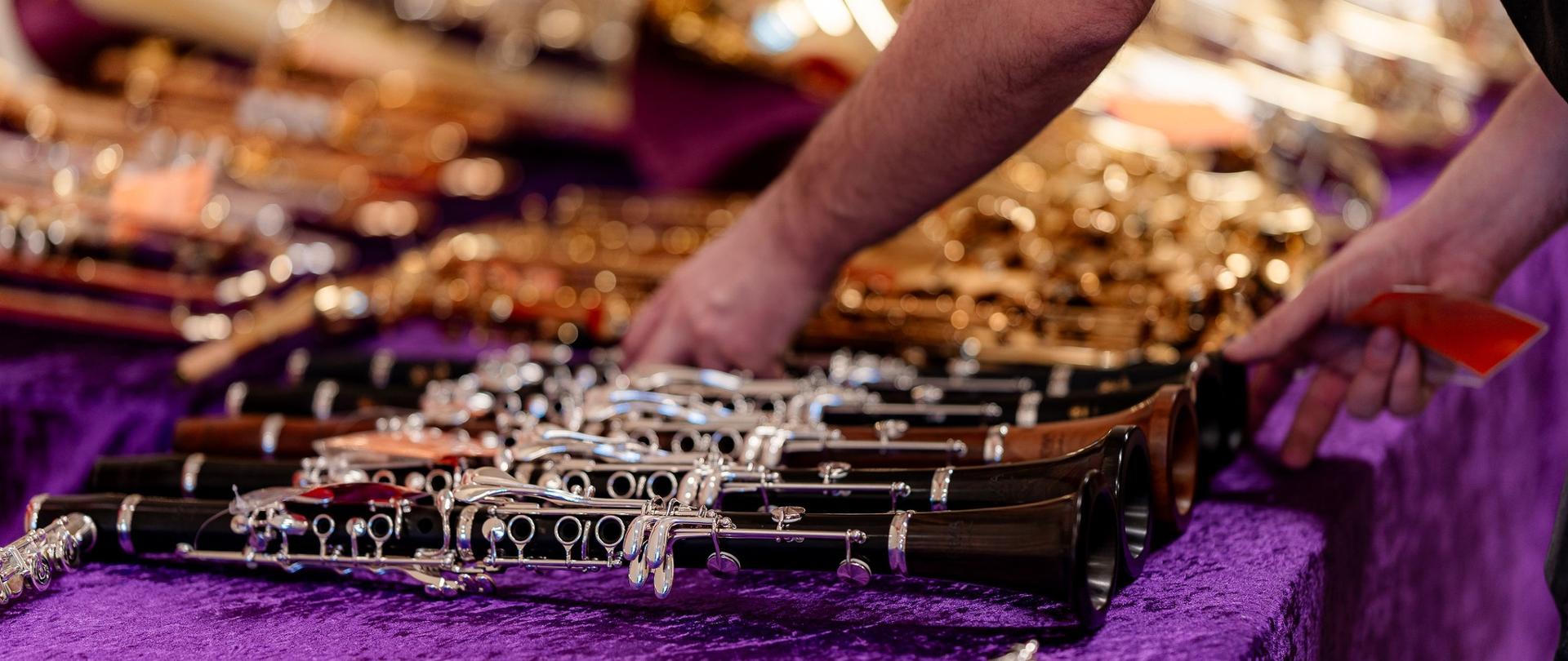 Zbliżenie na dłonie osoby układającej instrumenty dęte drewniane na stole przykrytym fioletowym, pluszowym materiałem. W pierwszym planie wyraźnie widać kilka czarnych klarnetów ze srebrnymi klapami.