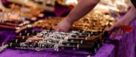 Zbliżenie na dłonie osoby układającej instrumenty dęte drewniane na stole przykrytym fioletowym, pluszowym materiałem. W pierwszym planie wyraźnie widać kilka czarnych klarnetów ze srebrnymi klapami.