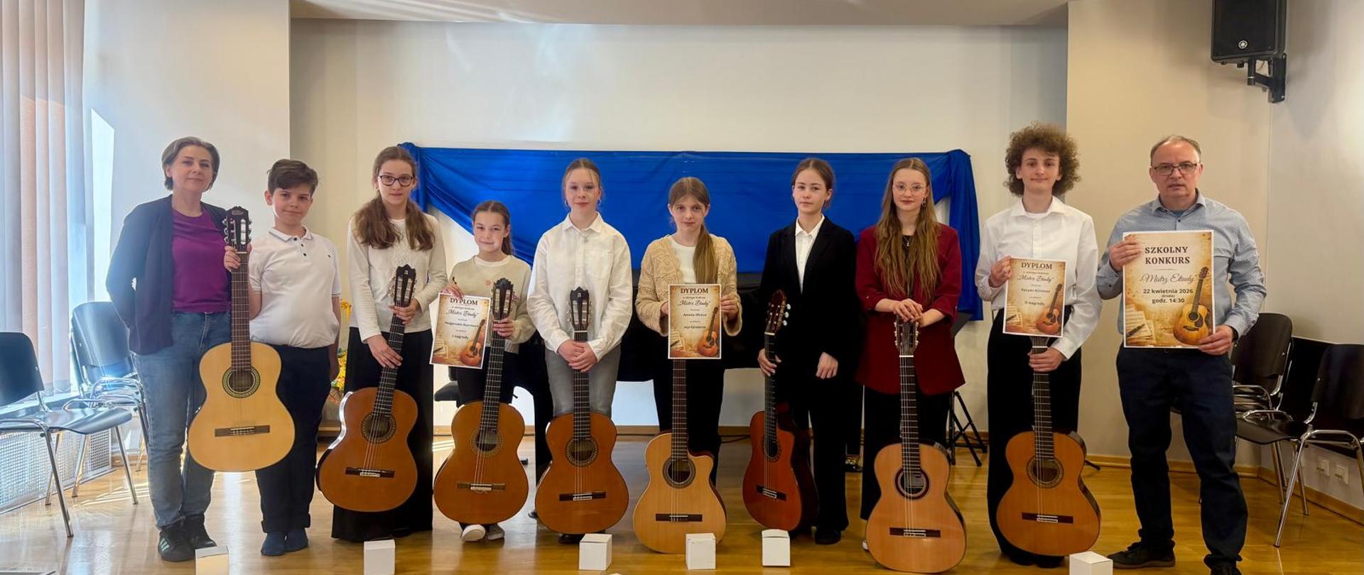Na scenie grupa uczniów. Każdy z nich trzyma gitarę. Trzy osoby trzymają dyplomy. Po prawej stronie mężczyzna w okularach i błękitnej koszuli, który trzyma plakat gitarowy. Po lewej stronie kobieta w jeansach, fioletowej bluzce i granatowym sweterku. W tle niebieski materiał.