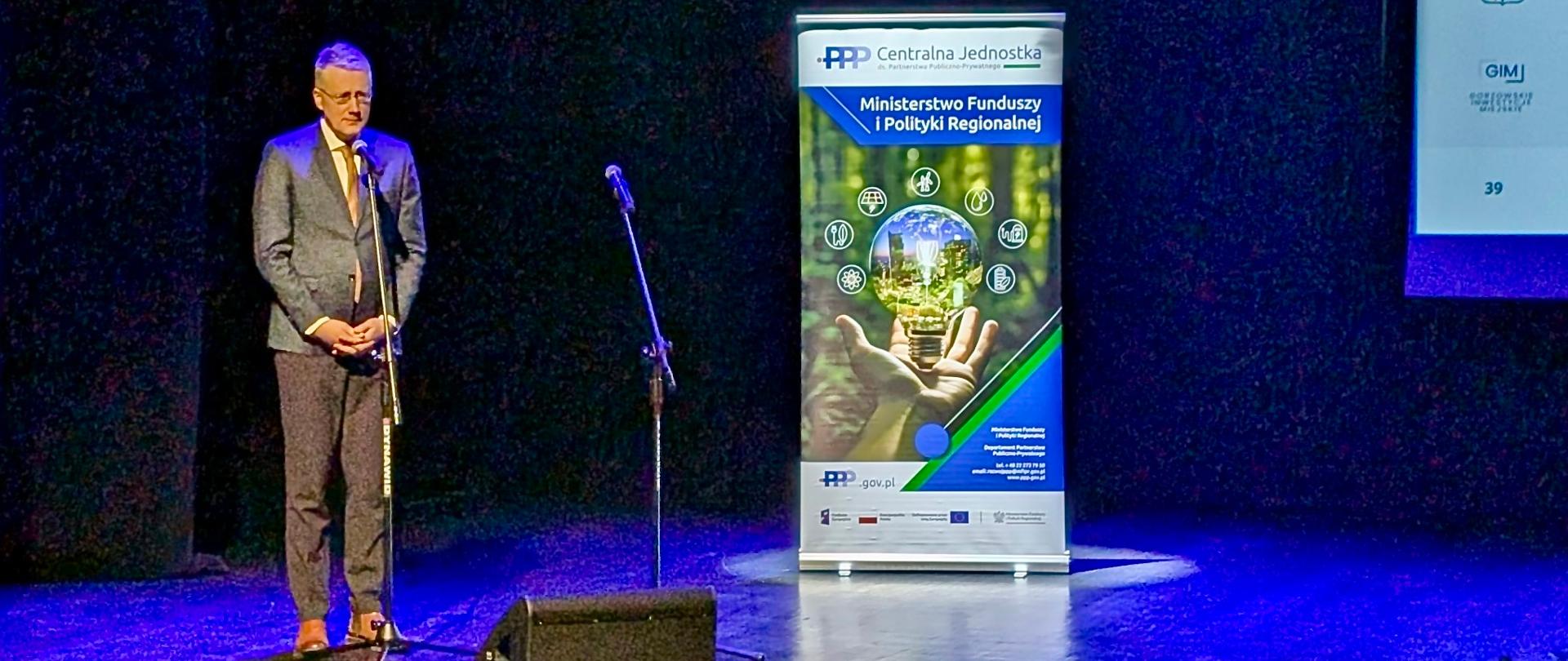 Na zdjęciu uczestnik konferencji stoi na scenie przed mikrofonem