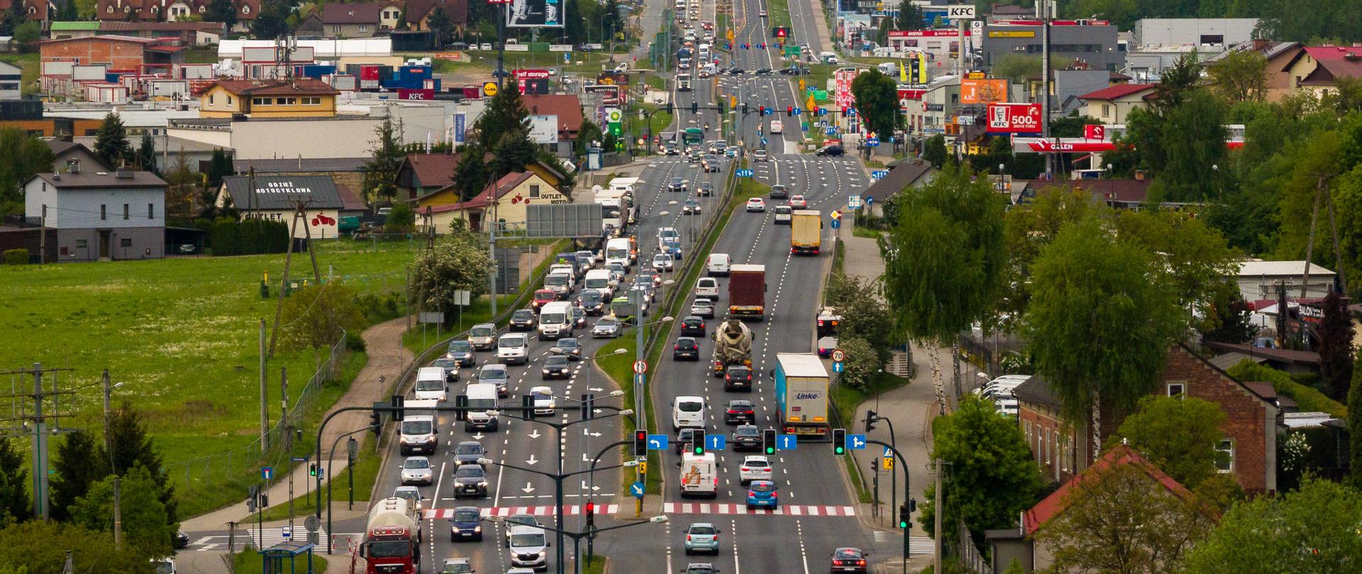 Widok na szeroką, wielopasmową drogę miejską z dużym natężeniem ruchu, otoczoną zabudową usługową i mieszkaniową, z zielenią w tle oraz pagórkowatym krajobrazem.