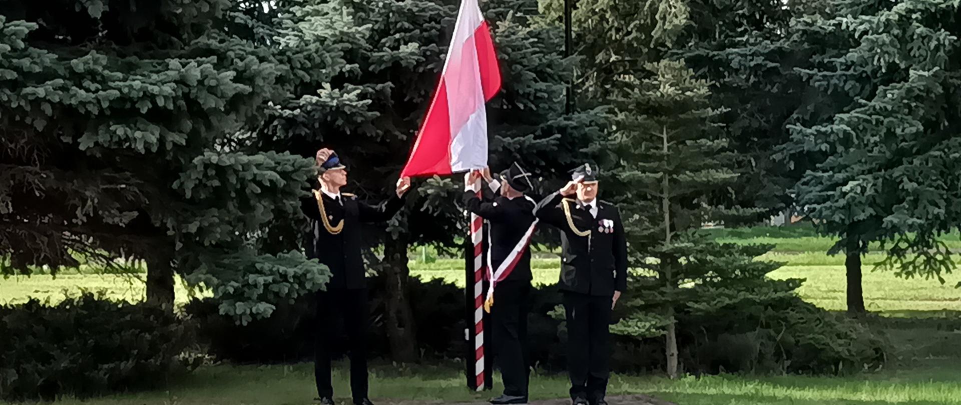 3 strażaków wiesza i wciąga flagę Rzeczypospolitej Polskiej na maszt