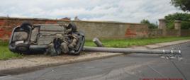 Kolizja w Trzeboszu. Auto osobowe uderzyło w słup energetyczny. Samochód leży na lewym boku. Obok na jezdni znajduje się złamany betonowy słup oraz zerwane przewody energetyczne. W tle murowane ogrodzenie gospodarstwa.
