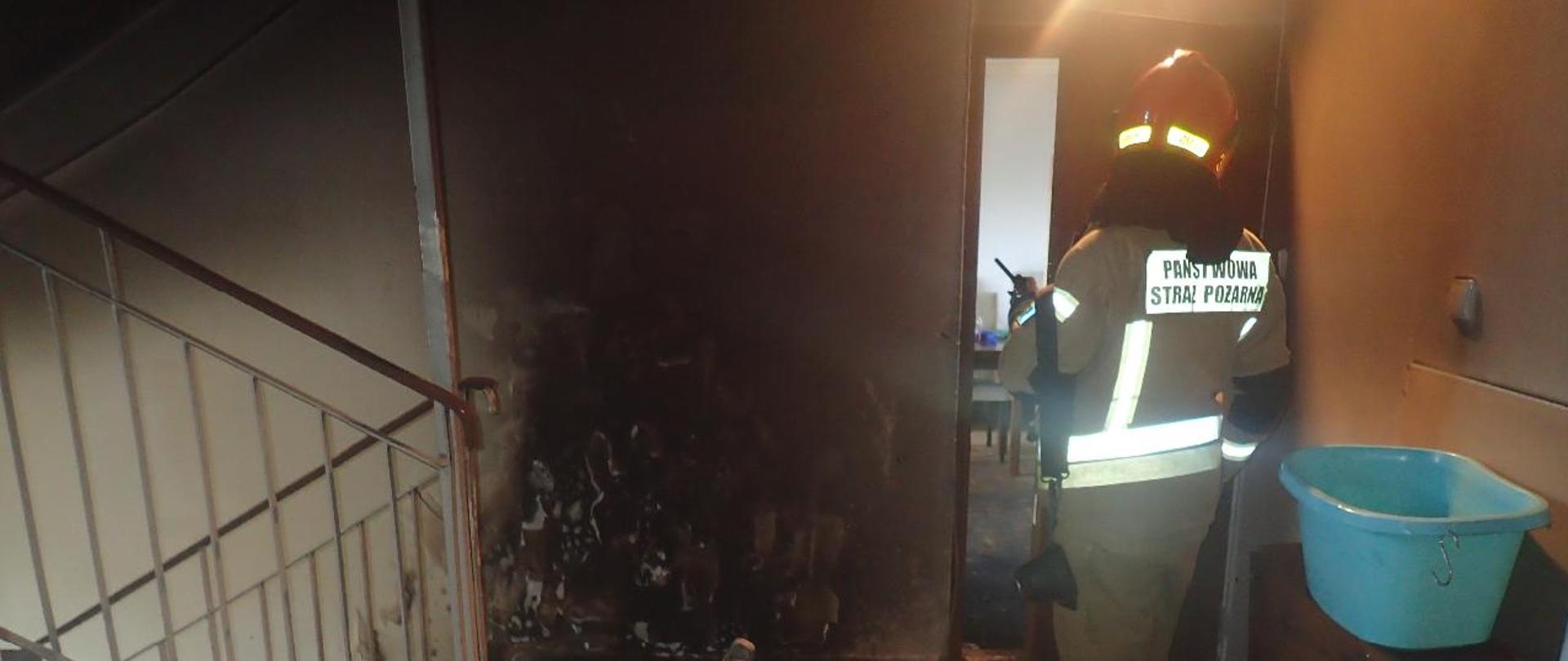 Zdjęcie 4 - na zdjęciu widać zgliszcza roweru elektrycznego i okopcone ściany i sufit na klatce schodowej oraz pracującego strażaka