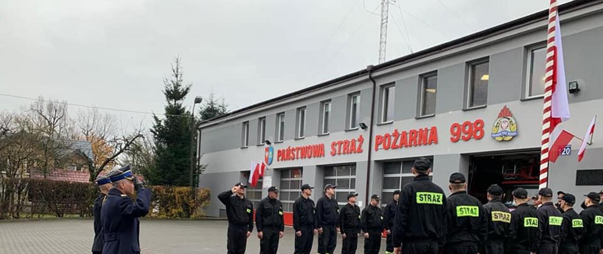 2 pododdziały strażaków stoją naprzeciwko masztu flagowego - oddają honory podczas odgrywania hymnu państwowego