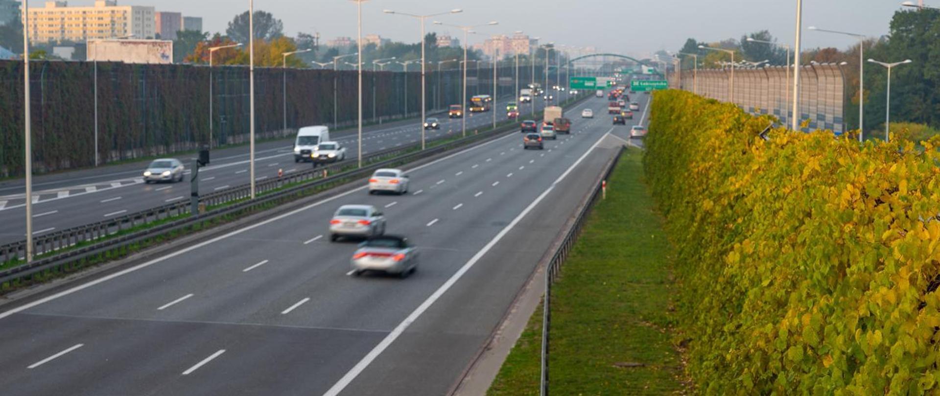 Pojazdy poruszające się drogą ekspresową. Pochmurny, jesienny dzień. Po obu stronach drogi stoją ekrany akustyczne. W oddali wiadukt.