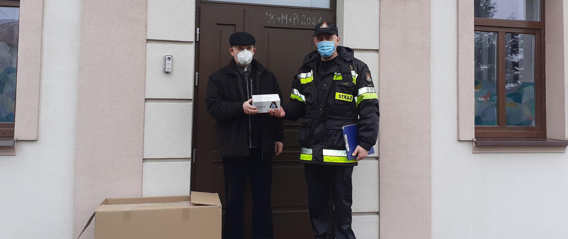 Widać strażaka i księdza. Ksiądz trzyma w ręku paczkę maseczek. Na dalszym planie widać część budynku. Po lewej stronie księdza stoi karton, za nim znajdują się drzwi do budynku.
