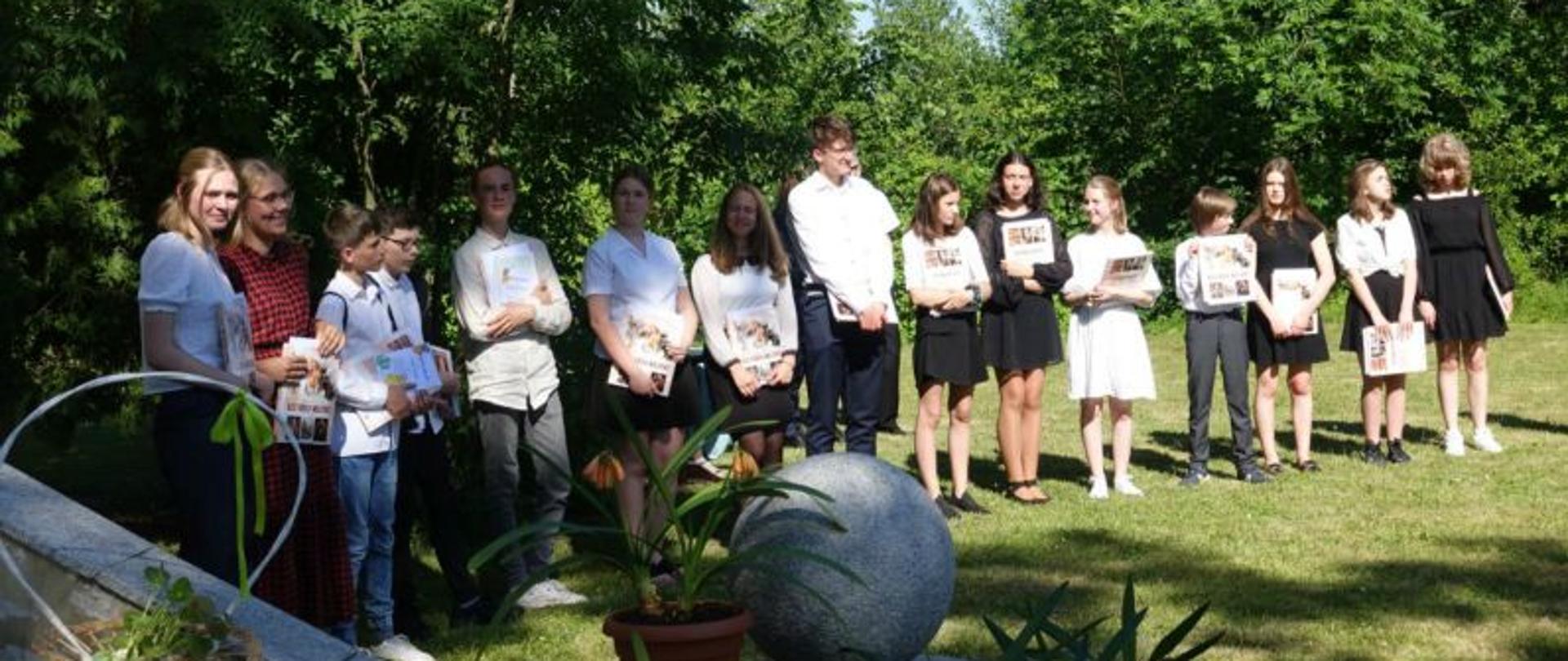Zdjęcie przedstawia grupę trzynastu uczniów stojących na trawniku za budynkiem A PSM w Słupsku. Uczniowie w strojach galowych trzymają dyplomy i świadectwa.