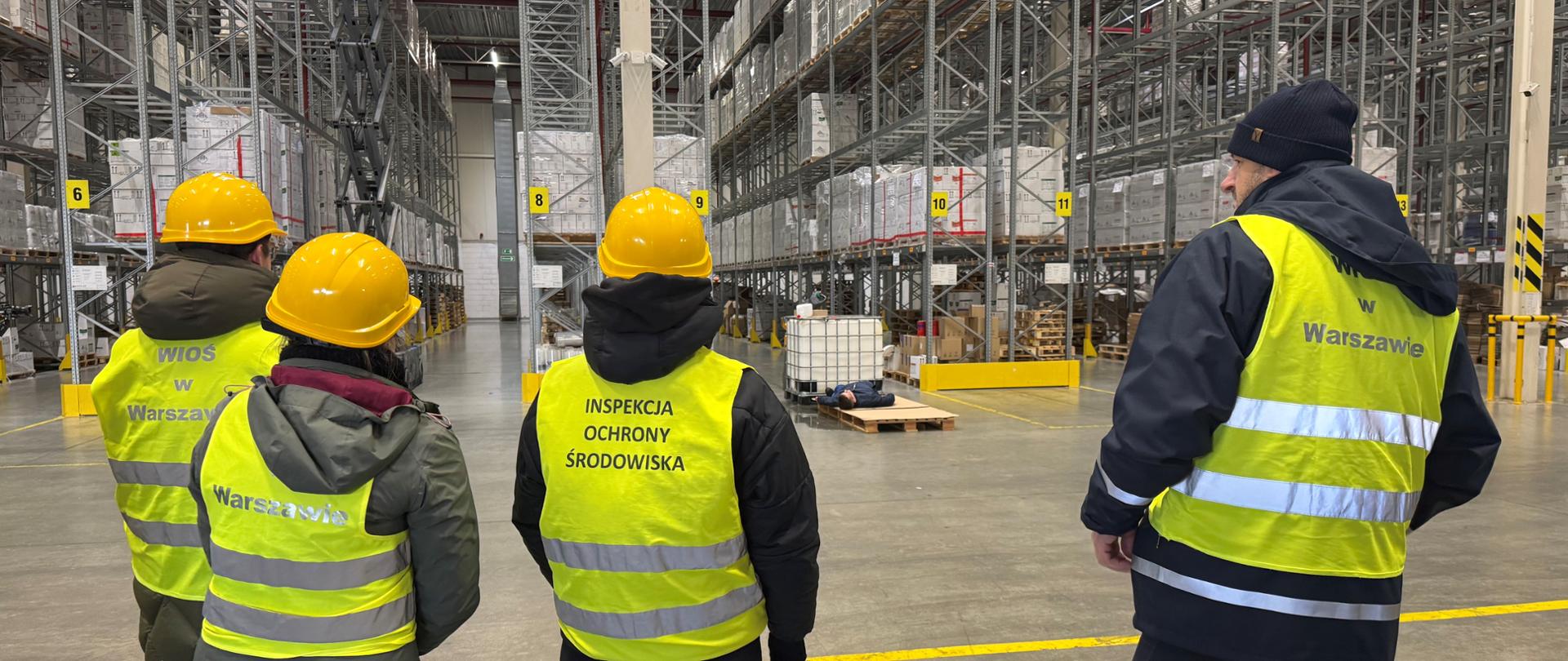 Czworo inspektorów Inspektoratu Ochrony Środowiska w Warszawie obserwuje ćwiczenia w dużej hali magazynowej W tle widoczne są wysokie regały z towarem i stojący na betonowej podłodze biały, plastikowy pojemnik o pojemności 1000 l.