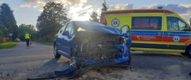 Widać mocno uszkodzony samochód osobowy od przodu, jest na drodze, po prawej karetka pogotowia