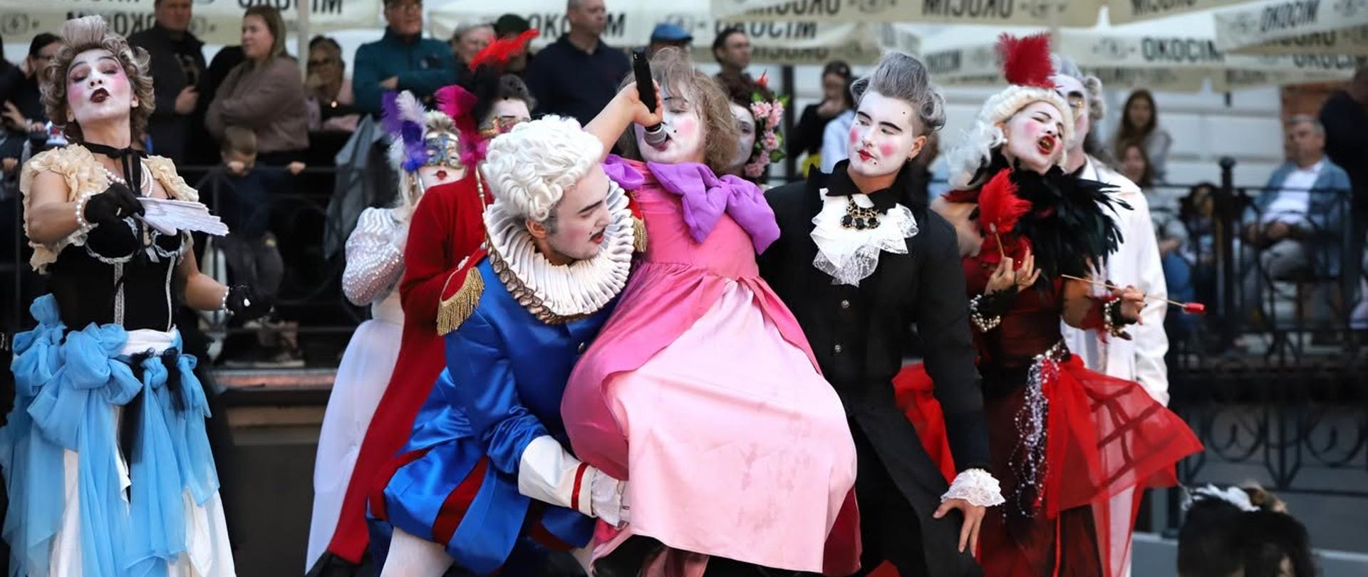 Grupa performerów w barokowych kostiumach i scenicznych makijażach występuje przed publicznością podczas festiwalu LA STRADA. W centrum kadru jedna z osób w niebieskim stroju podtrzymuje postać w różowej sukni, a wokół nich pozostali wykonawcy tworzą ekspresyjną, teatralną scenę.