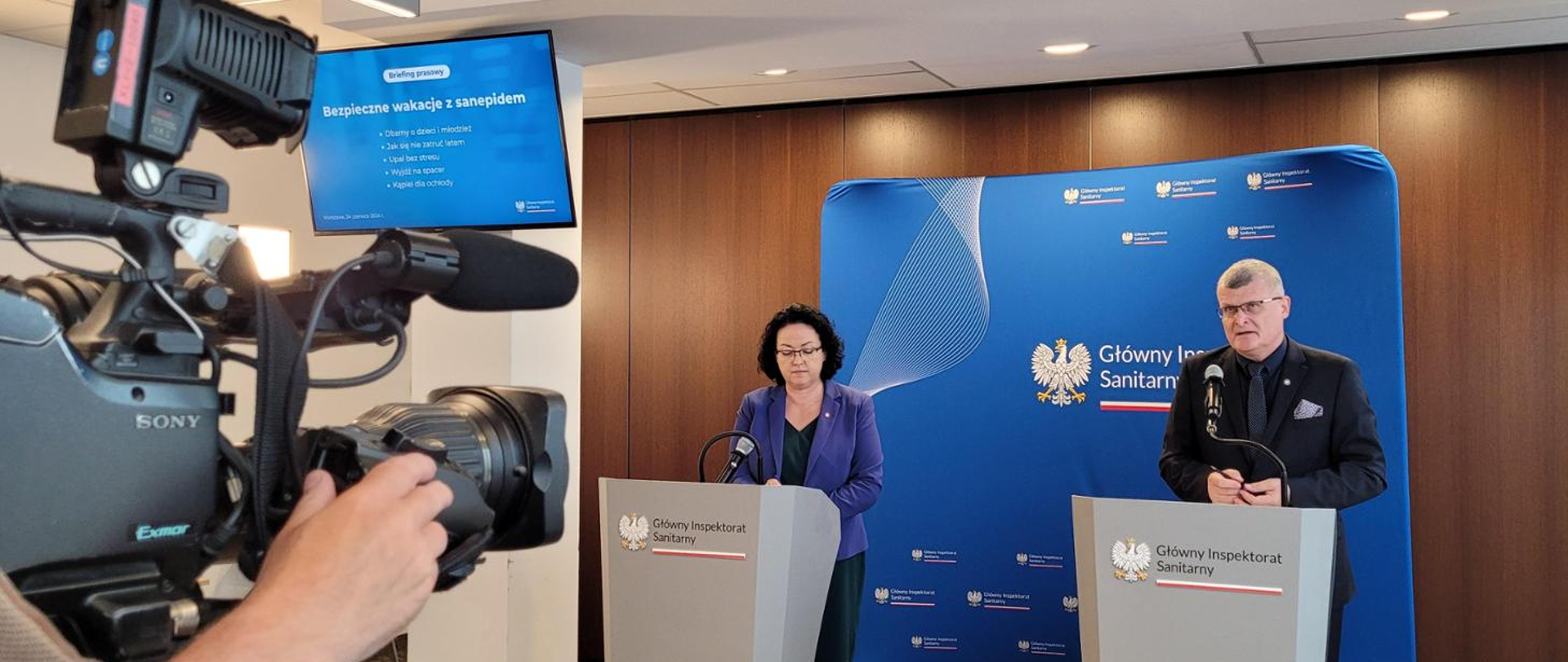 Na obrazku widzimy konferencję prasową, na której przemawia dwoje ludzi. Po lewej stronie stoi kobieta w ciemnofioletowej marynarce, a po prawej mężczyzna w ciemnym garniturze. Oboje stoją za mównicami oznaczonymi logo i nazwą "Główny Inspektorat Sanitarny". Na pierwszym planie z lewej strony znajduje się kamera, na której widać ręce operatora.