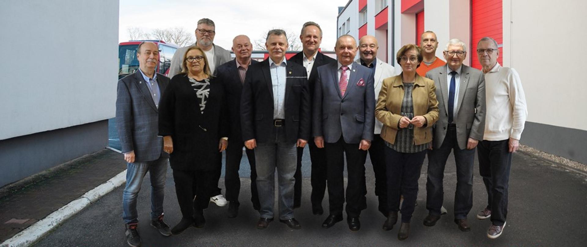 Dwanaście osób stojących obok siebie. W tle widać budynek z bramami garażowymi i autobus