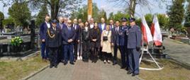 Rawicki cmentarz parafialny. Zdjęcie grupowe wszystkich uczestników uroczystości przed krzyżem katyńskim. Wśród uczestników są przedstawiciele służb mundurowych: PSP, Policji i Służby Więziennej. Z prawej strony stoi stojak z biało-zerwonymi flagami.W tle drzewa i nagrobki.