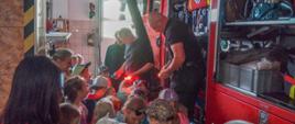 Zdjęcie przedstawia grupę dzieci podczas zapoznania ze sprzętem strażackim w pomieszczeniu garażu na tle pojazdów strażackich koloru czerwonego.