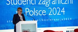 Za mównicą stoi wiceminister Mrówczyńska i mówi do mikrofonu, za nią na ścianie ekran z napisem XVII konferencja - Studenci zagraniczni w Polsce 2024.
