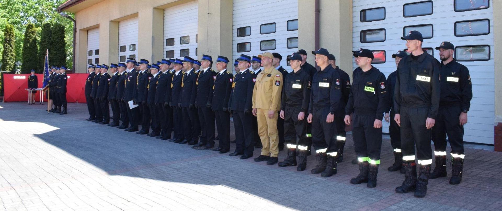 Na zdjęciu widzimy strażaków ustawionych w dwuszeregu, część z nich ubrana jest w umundurowanie galowe, a część w koszarowe. Za nimi stoi poczet sztandarowy. W tle widać budynek strażnicy.