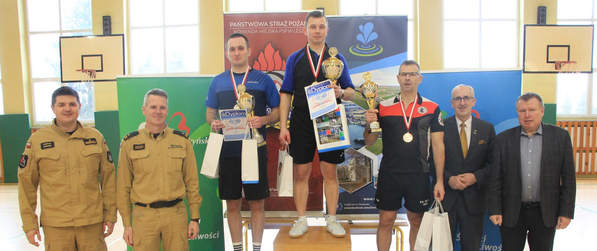 na podium stoją sportowcy z dyplomami w reku oraz papierowymi siatkami, obok nich z lewej strony stoi dwóch strażaków w miodowych mundurach a z prawej dwóch mężczyzn w garniturach