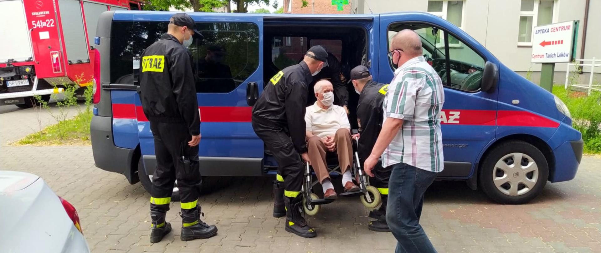 Zdjęcie przedstawia czterech strażaków pomagających osobie na wózku inwalidzkim udać się na szczepienie przeciwko Covid-19.