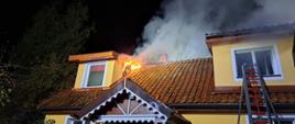 Zdjęcie przedstawia budynek mieszkalny jednorodzinny objęty pożarem. Widoczny jest wydobywający się dym i płomienie z dachu obiektu.