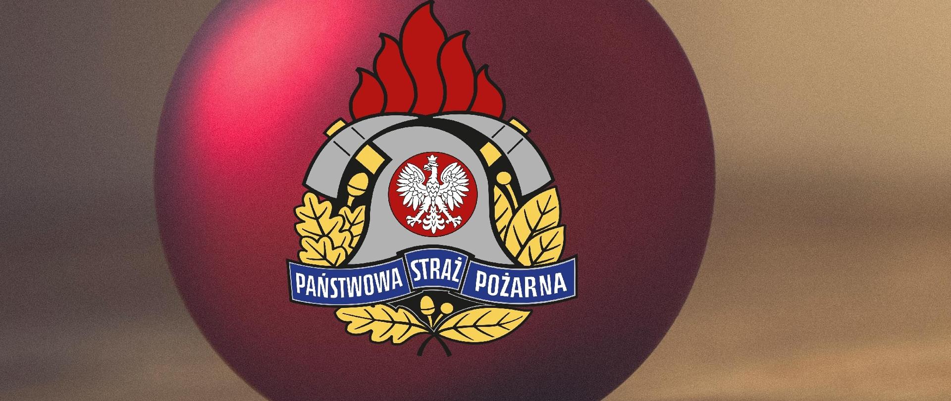 Zdjęcie przedstawia logotyp Państwowej Straży Pożarnej umoszczony na czerwonej bombce choinkowej. Bombka leży pionowo do góry na brązowej powierzchni o strukturze drewna. Ściana za bombką jest rozmyta również w jasnobrązowym kolorze. Po prawej stronie od bombki znajdują się życzenia koloru czarnego. 