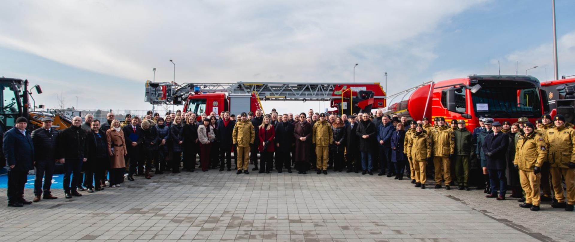 Duża grupa strażaków, przedstawicieli władz i gości pozuje do wspólnego zdjęcia przed szeregiem nowych pojazdów ratowniczych. Za nimi stoją wozy bojowe, drabiny i sprzęt specjalistyczny przekazany jednostkom PSP podczas wydarzenia na ITAKA ARENA.