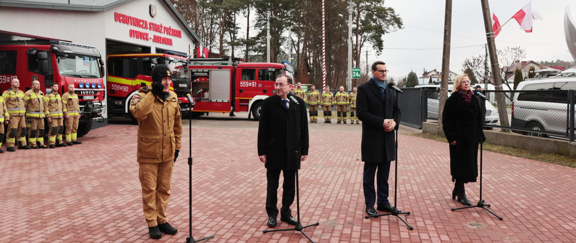 Konferencja prasowa. Premier, Ministrowie oraz Komendant Główny stojący przy mikrofonach. W tle remiza oraz samochody i druhowie ochotniczej straży pożarnej