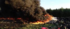 Zdjęcie przedstawia pożar składowiska opon. Widoczne są palące się opony i kłęby czarnego dymu. Po prawej stronie strażacy gaszący pożar.