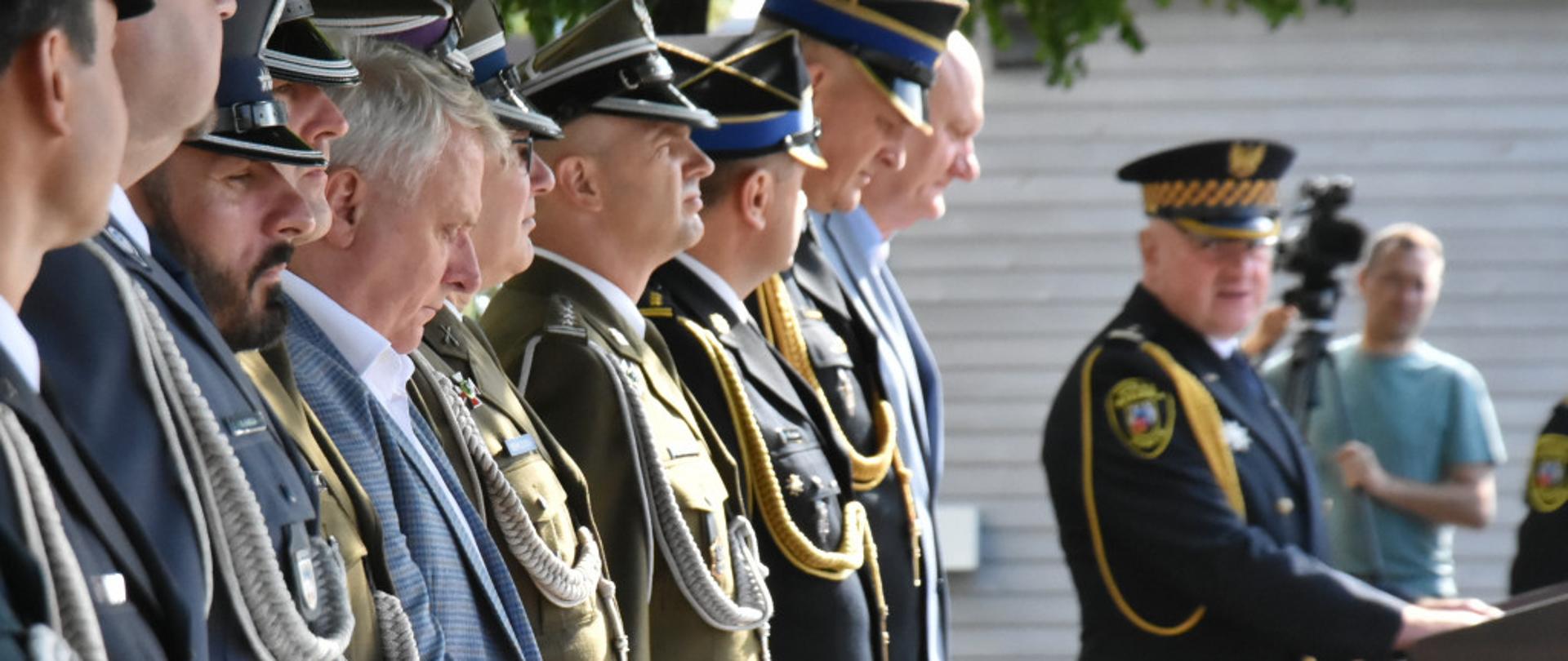 W szeregu stoją goście uroczystości, w tym komendanci PSP. W tle, przy mównicy stoi Komendant Straży Miejskiej i wzwraca się do gości. Na dalszym planie operator z kamerą.