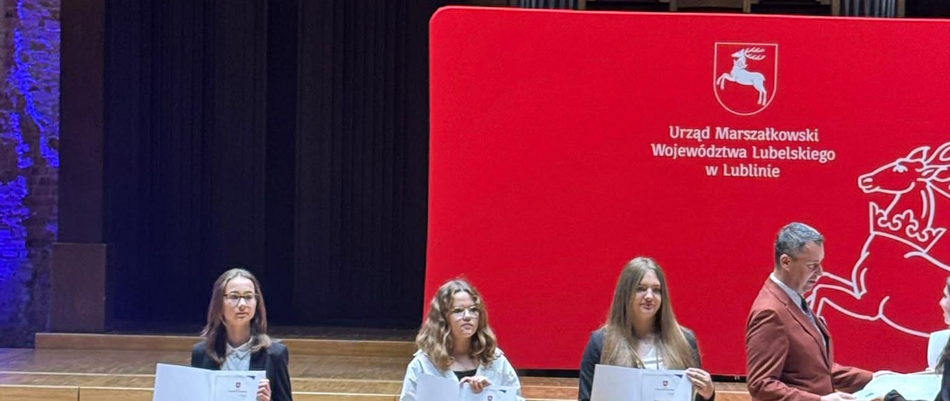 Dziewczynka elegancko ubrana stoi z dyplomem w ręku