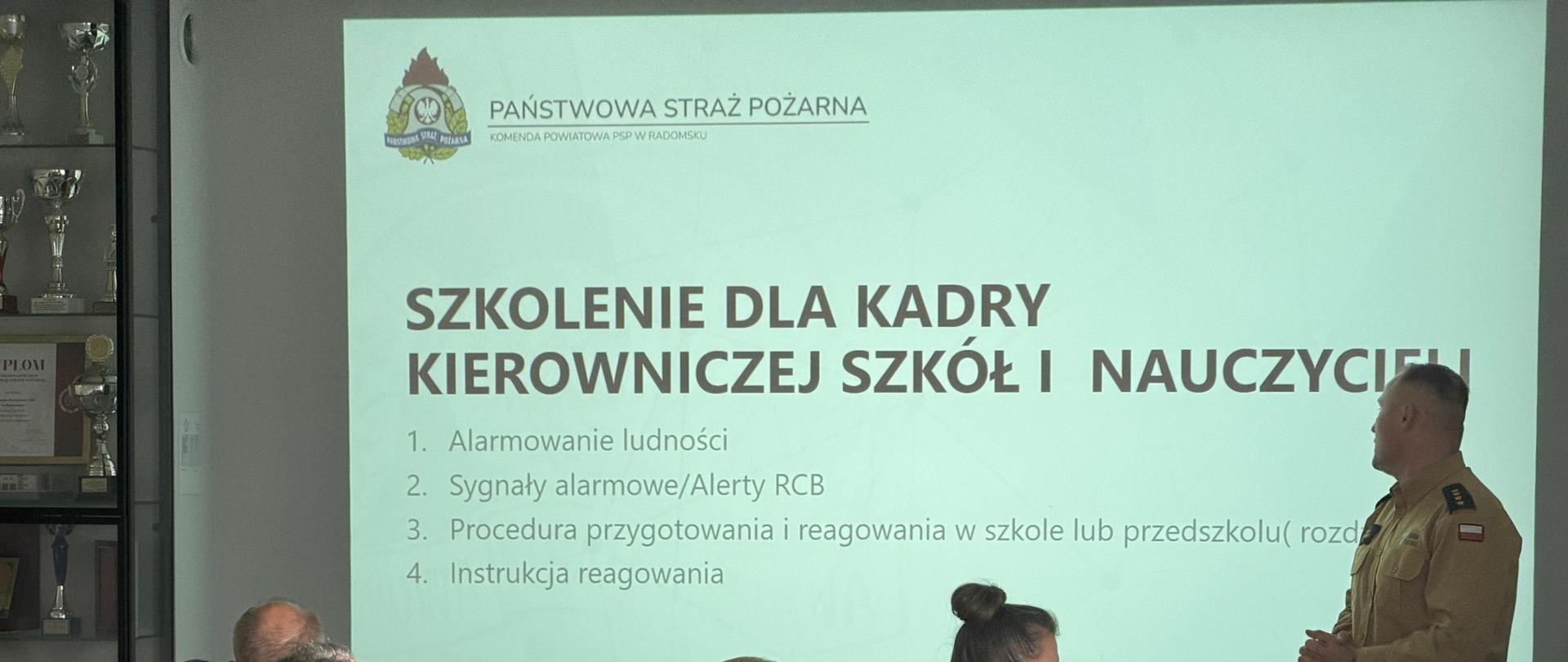 Funkcjonariusz PSP w piaskowym mundurze prowadzi szkolenie dla przedstawicieli placówek oświatowych. Na ekranie widoczna jest prezentacja dotycząca sygnałów alarmowych, alertów RCB oraz zasad postępowania w szkołach i przedszkolach.