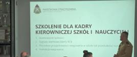 Funkcjonariusz PSP w piaskowym mundurze prowadzi szkolenie dla przedstawicieli placówek oświatowych. Na ekranie widoczna jest prezentacja dotycząca sygnałów alarmowych, alertów RCB oraz zasad postępowania w szkołach i przedszkolach.
