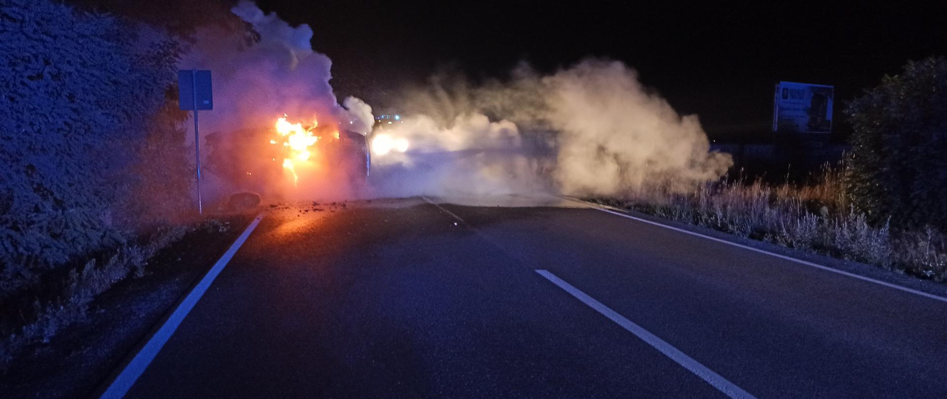 Na zdjęciu widać pożar samochodu osobowego. Panuje silne zadymienie oraz płomienie. Samochód leży na boku na jezdni. Pora nocna. 