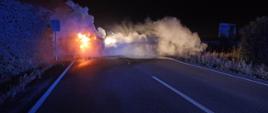 Na zdjęciu widać pożar samochodu osobowego. Panuje silne zadymienie oraz płomienie. Samochód leży na boku na jezdni. Pora nocna. 